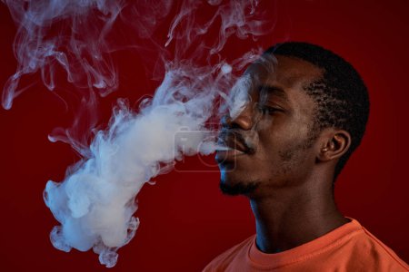 Foto de Joven hombre negro con camiseta naranja fumando cigarrillo electrónico frente a la cámara sobre fondo rojo y nube exhaladora de humo blanco - Imagen libre de derechos