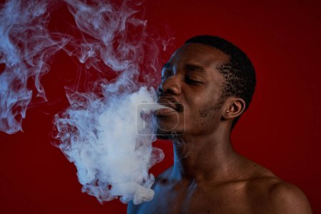 Téléchargez les photos : Shirtless Afro-Américain gars soufflant nuage de fumée épaisse tout en se tenant dans l'isolement devant la caméra sur fond rouge - en image libre de droit