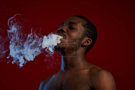 Foto de Acercador masculino afroamericano sin camisa que libera una espesa nube de humo de su boca mientras disfruta fumando cigarrillo electrónico - Imagen libre de derechos