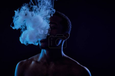 Téléchargez les photos : Jeune homme afro-américain debout devant la caméra dans l'obscurité et soufflant un nuage de vapeur hors de sa bouche tout en fumant - en image libre de droit