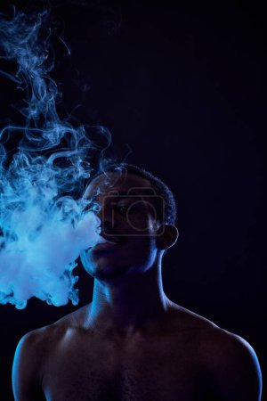 Téléchargez les photos : Jeune homme debout devant la caméra dans l'obscurité et libérant de la vapeur de sa bouche pendant le processus de fumer e-cigarette - en image libre de droit