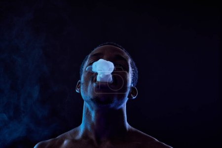 Foto de Vista frontal de un joven negro sin camisa levantando la cabeza mientras fuma cigarrillo electrónico o narguile mientras está de pie en la oscuridad - Imagen libre de derechos