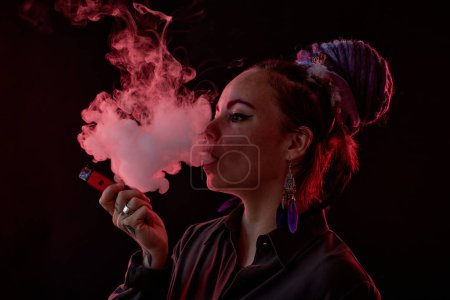 Photo for Young woman in casualwear holding electronic cigarette and blowing smoke out of her mouth while standing in darkness - Royalty Free Image
