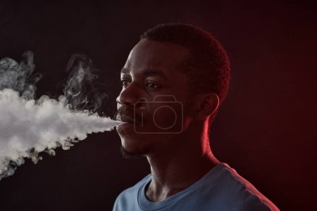 Téléchargez les photos : Jeune homme afro-américain expirant nuage blanc de fumée épaisse tout en se tenant devant la caméra dans la pièce sombre dans l'isolement - en image libre de droit