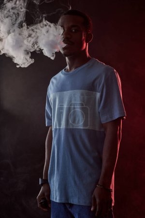 Foto de Joven hombre afroamericano en camiseta y jeans sosteniendo cigarrillo electrónico mientras está parado frente a la cámara y fumando en la oscuridad - Imagen libre de derechos