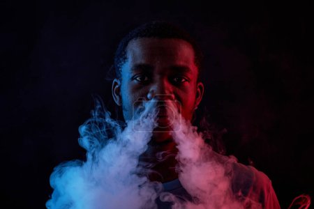 Foto de Retrato de cerca del joven afroamericano mirando a la cámara mientras está de pie en la oscuridad y soplando humo de cigarrillo de las fosas nasales - Imagen libre de derechos
