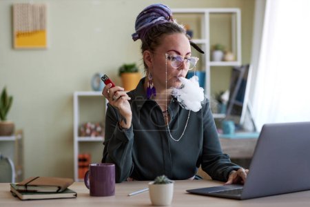 Téléchargez les photos : Jeune femme d'affaires avec cigarette électronique assise devant un ordinateur portable, regardant l'écran et libérant un nuage de fumée de la bouche et des narines - en image libre de droit