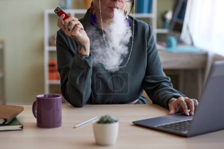 Téléchargez les photos : Gros plan d'une jeune créatrice qui fume de la cigarette électronique sur son lieu de travail tout en étant assise devant un ordinateur portable et en réseautant - en image libre de droit