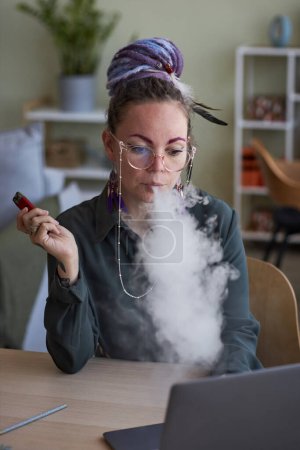 Photo for Young female manager in eyeglasses and casualwear smoking electronic cigarette in front of laptop during work over business project - Royalty Free Image