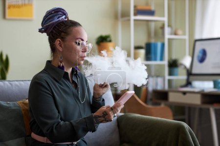 Foto de Joven mujer de negocios desplazándose en el teléfono móvil y fumar cigarrillo electrónico mientras está sentado en el sofá en la oficina en casa en el descanso - Imagen libre de derechos