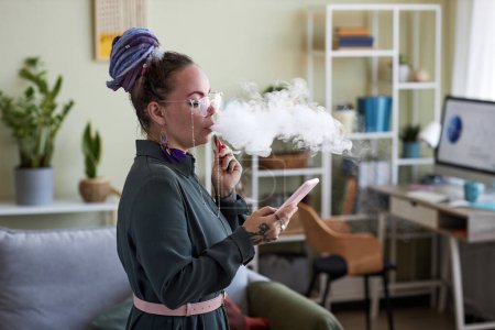 Photo for Young businesswoman smoking electronic cigarette and texting in mobile phone while standing in living room in home office at break - Royalty Free Image
