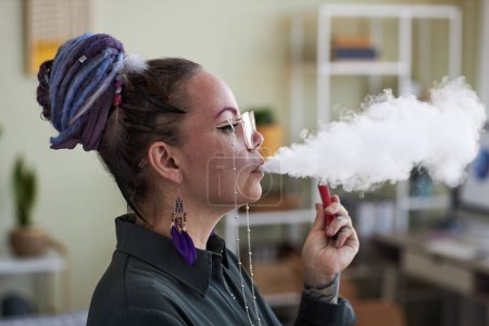 Téléchargez les photos : Vue latérale de la jeune femme en lunettes soufflant un nuage blanc de fumée épaisse hors de sa bouche tout en fumant devant la caméra - en image libre de droit