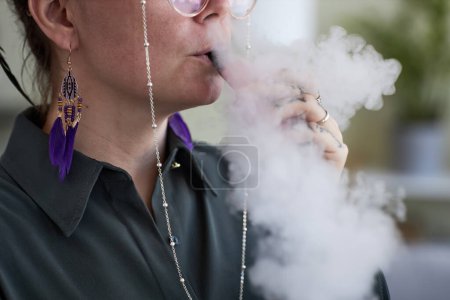 Photo for Close-up of young female smoker releasing cloud of white thick vapor while keeping electronic cigarette in her mouth during smoking - Royalty Free Image