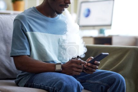 Photo for Cropped shot of African American guy with vape pen using mobile phone while sitting on couch in home office during break and relaxing - Royalty Free Image