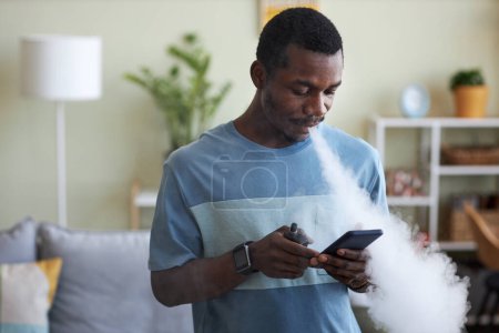 Photo for Young African American businessman scrolling in smartphone and smoking electronic cigarette while standing in front of camera at home - Royalty Free Image
