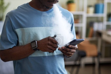 Foto de Chico afroamericano con e-cigarrillo en la mano mensajes de texto en el teléfono inteligente y la liberación de nubes de vapor grueso blanco de su boca - Imagen libre de derechos