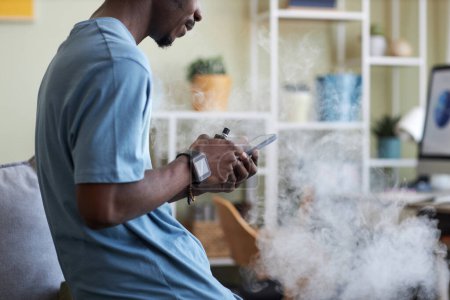 Téléchargez les photos : Vue latérale du jeune homme noir en t-shirt debout devant la caméra dans le salon et textos dans le smartphone tout en fumant e-cigarette - en image libre de droit