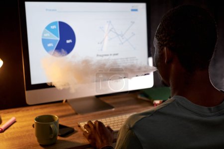 Téléchargez les photos : Jeune homme d'affaires fumant de la cigarette électronique assis devant un ordinateur avec des données graphiques à l'écran et travaillant tard dans la soirée - en image libre de droit