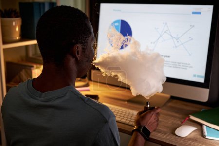 Photo for Young male analyst smoking electronic cigarette in front of computer screen by his workplace while analyzing graphic data - Royalty Free Image
