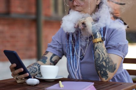Foto de Primer plano de la mujer joven con cigarrillo electrónico sentado a la mesa en la cafetería al aire libre, soplando vapor de su boca y desplazándose en el teléfono inteligente - Imagen libre de derechos