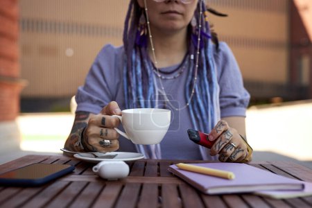 Photo for Close-up of young woman with cup of coffee and electronic cigarette sitting by table in outdoor cafe at break and enjoying rest - Royalty Free Image