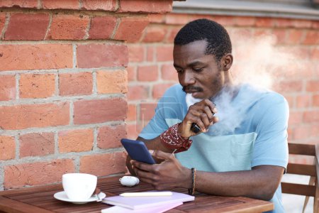 Photo for African American guy in blue t-shirt scrolling in smartphone and releasing cloud of vapor out of mouth while sitting by table in outdoor cafe - Royalty Free Image