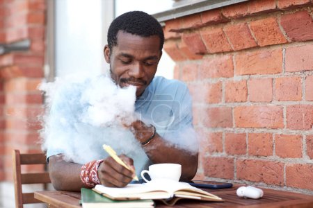 Téléchargez les photos : Jeune homme d'affaires afro-américain fumant une cigarette électronique et prenant des notes dans un cahier assis à table dans un café extérieur - en image libre de droit