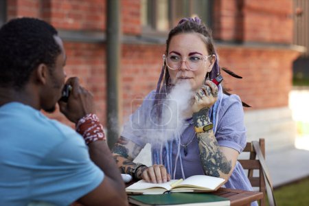 Photo for Young businesswoman looking at her colleague while both smoking vape pens and discussing working points at outdoor business meeting - Royalty Free Image