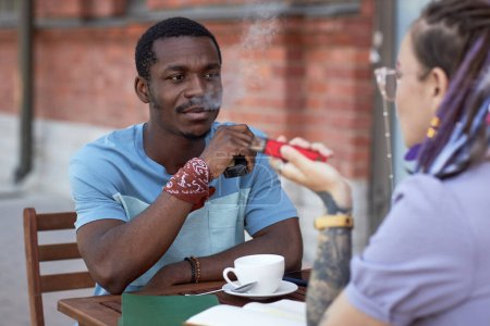 Foto de Joven hombre de negocios afroamericano mirando a una colega durante la comunicación mientras ambos fuman cigarrillos electrónicos en la reunión - Imagen libre de derechos