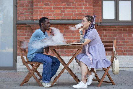 Photo for Side view of two young intercultural colleagues smoking electronic cigarettes while sitting by table and organizing work at meeting - Royalty Free Image