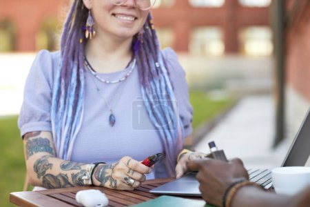 Photo for Close-up of young smiling businesswoman with vape pen networking at meeting with African American male colleague in outdoor cafe - Royalty Free Image