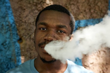 Foto de Rostro de un joven afroamericano que libera una nube de vapor blanco y espeso de su boca mientras fuma cigarrillos electrónicos y mira a la cámara - Imagen libre de derechos