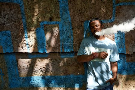 Foto de Chico afroamericano en azul camiseta fumar vapor pluma en día soleado mientras de pie contra la pared de la construcción en la sombra y mirando a la cámara - Imagen libre de derechos