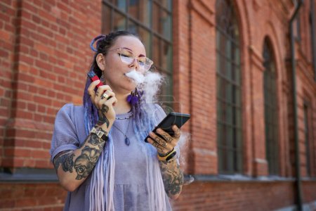 Photo for Young woman with tattoos on arms and hands smoking electronic cigarette and scrolling in mobile phone in urban environment - Royalty Free Image