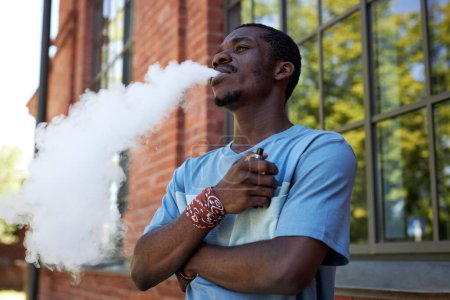 Foto de Joven hombre negro en camiseta azul claro fumar cigarrillo electrónico, mientras que de pie delante de la cámara contra el exterior del edificio moderno - Imagen libre de derechos