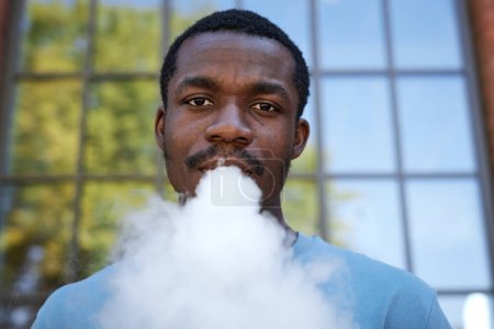 Photo for Close-up portrait of young African American man smoking electronic cigarette and looking at camera while blowing steam out of mouth - Royalty Free Image