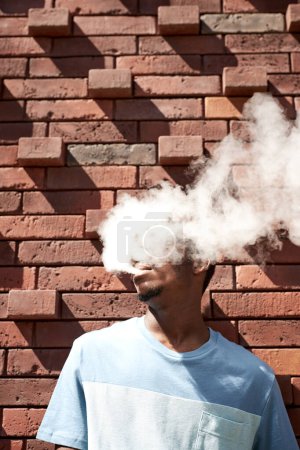 Foto de Joven parado junto a la pared de ladrillo rojo en un día soleado y soplando una nube de humo blanco y espeso de su boca mientras fuma cigarrillo electrónico - Imagen libre de derechos