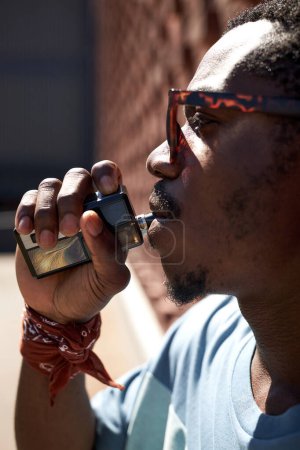Photo for Close-up of young African American man with electronic cigarette in mouth standing in front of camera in urban environment - Royalty Free Image