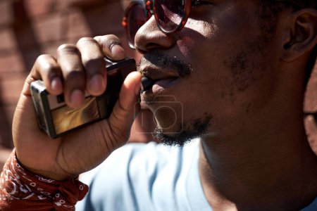 Photo for Close-up of young African American man in sunglasses holding electronic cigarette and keeping it in his mouth while smoking on sunny day - Royalty Free Image