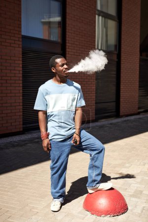Foto de Joven hombre negro en camiseta azul y jeans fumando en la calle en un día soleado mientras está de pie contra el exterior del edificio de ladrillo rojo - Imagen libre de derechos