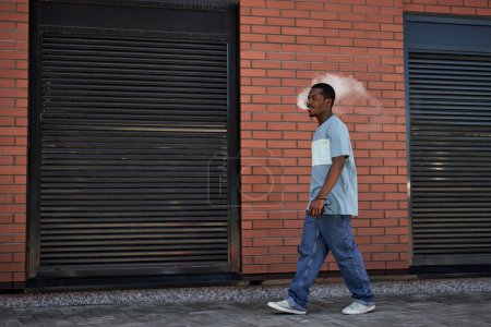 Foto de Vista lateral del joven hombre negro en ropa casual caminando a lo largo de la pared de ladrillo de la construcción y soplando nube de vapor de su boca - Imagen libre de derechos