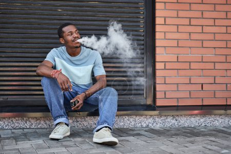 Foto de Chico afroamericano en ropa de casualwear fumar cigarrillo electrónico al aire libre mientras está sentado en la acera contra la pared de ladrillo rojo del edificio - Imagen libre de derechos
