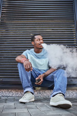 Foto de Joven hombre afroamericano descansado en ropa casual sentado en la acera por la pared del edificio moderno y fumar cigarrillo electrónico - Imagen libre de derechos