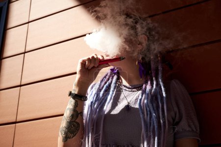 Photo for Hidden face of young woman with dreadlocks holding e-cigarette while standing against wall and blowing cloud of smoke out of mouth - Royalty Free Image
