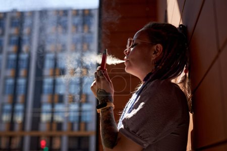 Photo for Young woman with vape pen in hand standing by wall of building and releasing cloud of vapor out of her mouth while smoking - Royalty Free Image