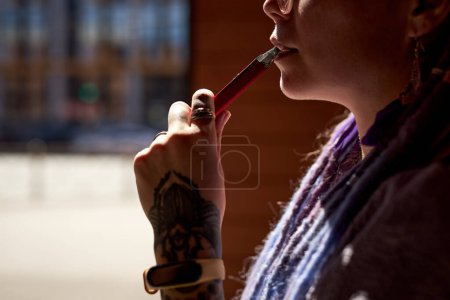 Téléchargez les photos : Gros plan d'une jeune femme tatouée tenant une cigarette électronique près de sa bouche devant une caméra extérieure - en image libre de droit