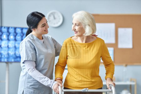 Photo for Waist up portrait of smiling young nurse helping senior woman using mobility walker in medical clinic - Royalty Free Image
