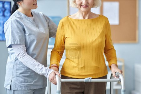 Téléchargez les photos : Portrait recadré d'une femme âgée utilisant une marchette de mobilité à la clinique avec une infirmière aidant - en image libre de droit