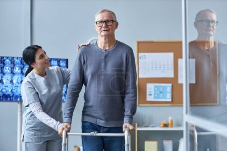 Téléchargez les photos : Portrait taille haute d'une infirmière souriante aidant un aîné à utiliser une marchette à mobilité réduite dans une maison de retraite - en image libre de droit