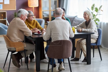 Téléchargez les photos : Groupe d'aînés assis à la table ensemble et souriant joyeusement dans la salle commune de la maison de retraite - en image libre de droit